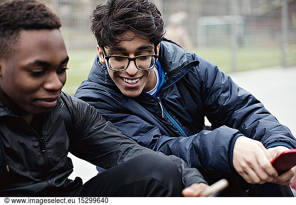 Smiling young man looking at friend's mobile phone against playing field in city during winter