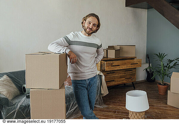 Smiling young man standing by cardboard boxes in living room