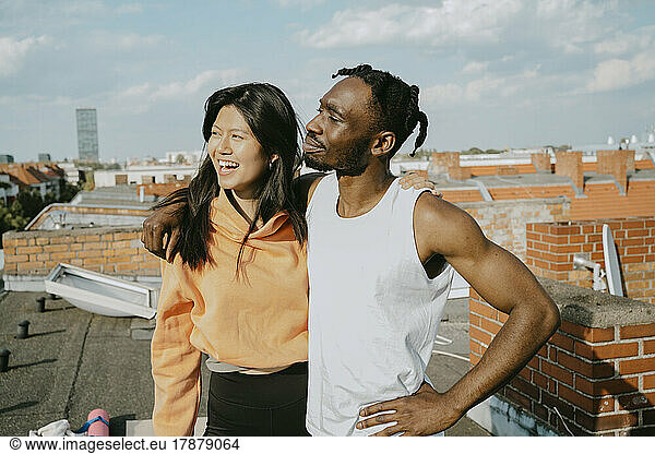 Smiling young man standing with arm around female friend looking away while standing on rooftop