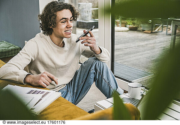Smiling young man talking on smart phone through speaker in living room