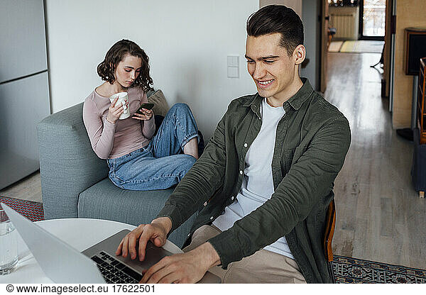Smiling young man using laptop by woman sitting with coffee cup and smart phone on sofa at home