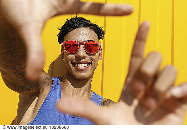 Smiling young man wearing red sunglasses gesturing finger frame leaning at yellow wall