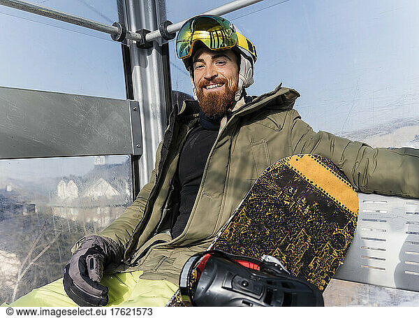 Smiling young man with snowboard sitting in cable car