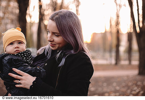 Smiling young mother carrying baby son standing in park in autumn