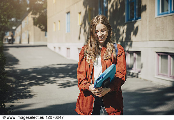 Smiling young student holding folder while standing on road in university campus