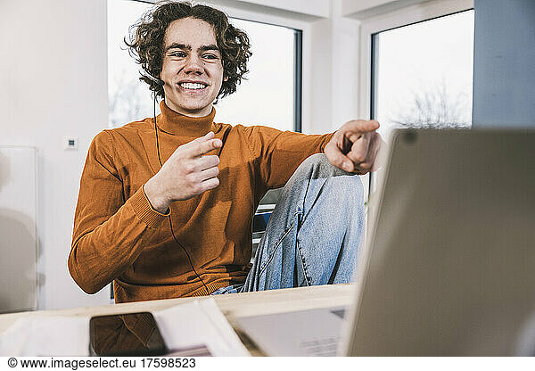 Smiling young student wearing headphones gesturing at table in living room