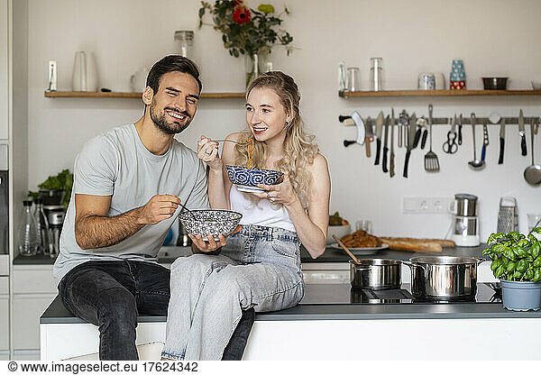 Smiling young woman and man eating spaghetti sitting on kitchen island at home
