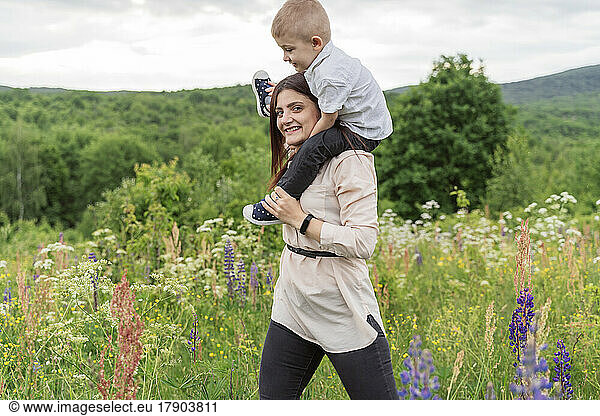 Smiling young woman carrying son on shoulders in meadow