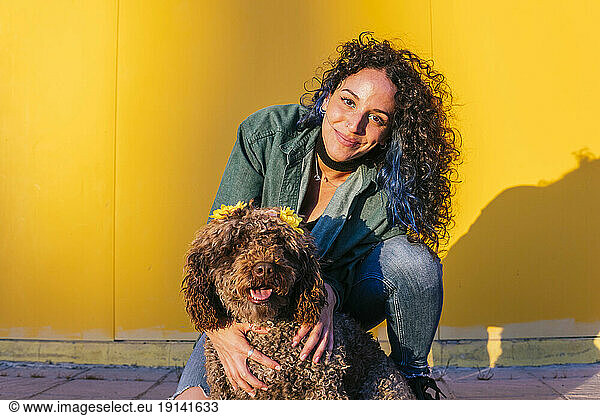 Smiling young woman crouching with water dog in front of yellow wall