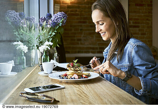 Smiling young woman eating pancakes in cafe