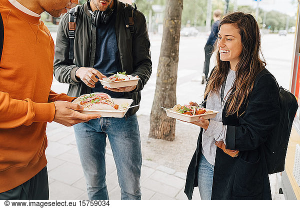 Smiling young woman enjoying meal with male friends while standing on street by food truck