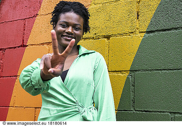 Smiling young woman gesturing peace sign in front of colorful wall