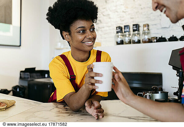 Smiling young woman giving disposable cup to colleague at checkout counter