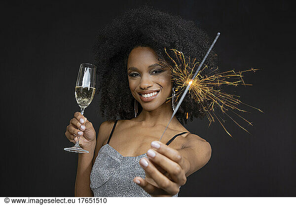 Smiling young woman holding champagne flute and sparkler against black background