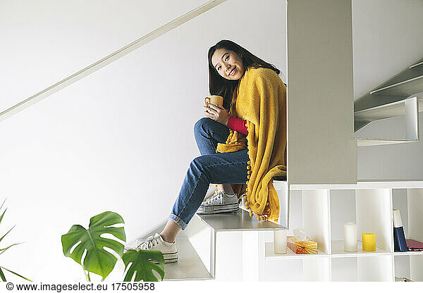 Smiling young woman holding coffee cup on steps at home
