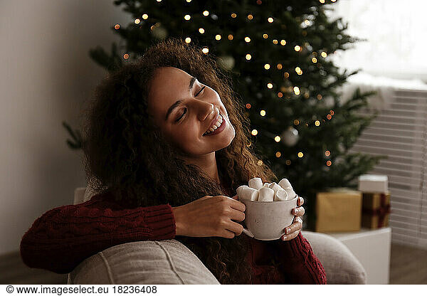 Smiling young woman holding cup of marshmallow cocoa sitting on sofa at home