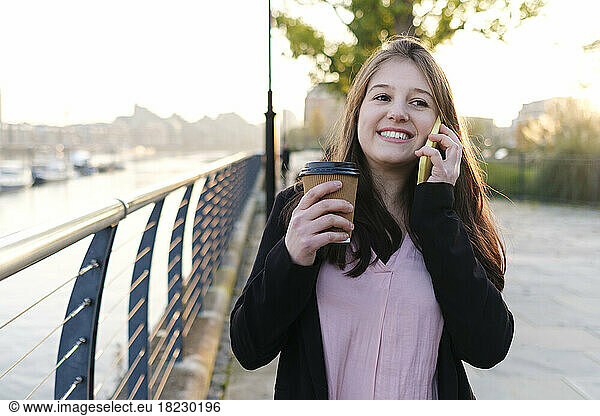 Smiling young woman holding disposable cup talking on phone by railing