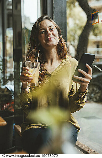 Smiling young woman holding glass of juice and smart phone in cafe