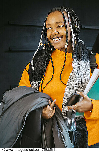 Smiling young woman holding jacket and books at community college