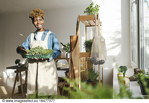 Smiling young woman holding microgreens container at home