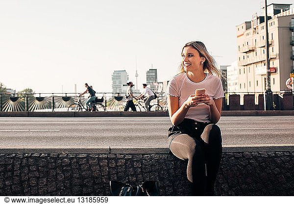 Smiling young woman holding mobile phone while sitting on retaining wall at street