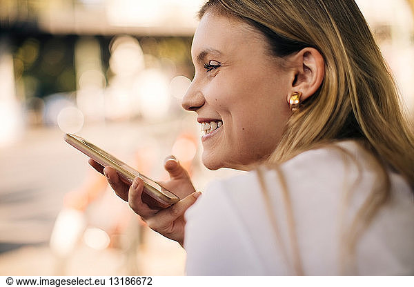 Smiling young woman holding mobile phone while sitting outdoors in city