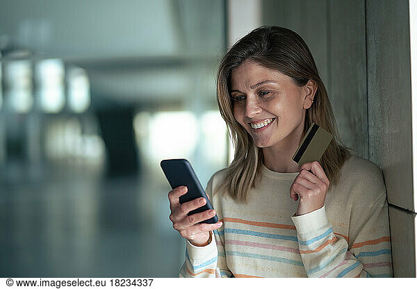 Smiling young woman holding smart phone and credit card leaning on wall