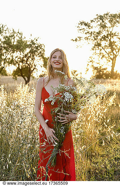 Smiling young woman holding wildflowers bouquet in field