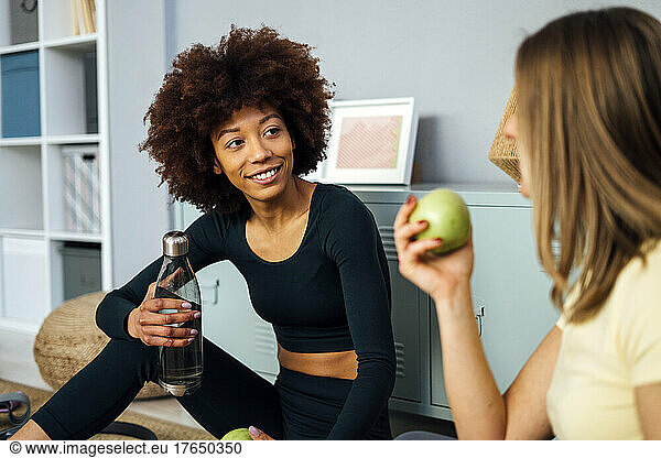 Smiling young woman holing water bottle looking at friend talking in living room