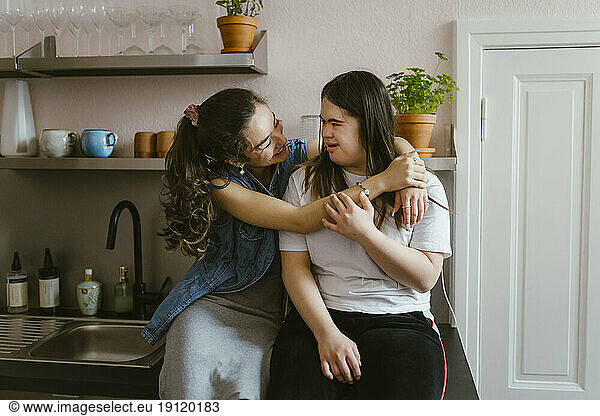 Smiling young woman hugging sister with down syndrome in kitchen at home