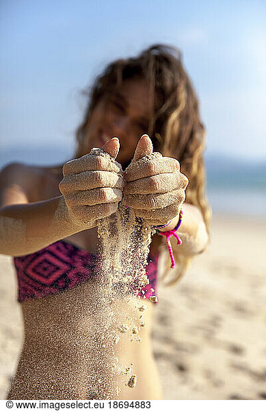 Smiling young woman in bikini pouring sand at beach