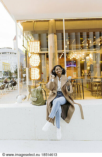 Smiling young woman listening to music through wireless headphones sitting in front of cafe