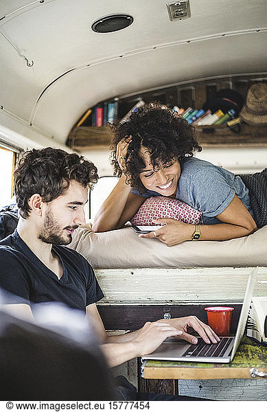 Smiling young woman lying on bed while looking at man using laptop in motor home