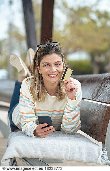Smiling young woman lying on bench holding credit card and smart phone