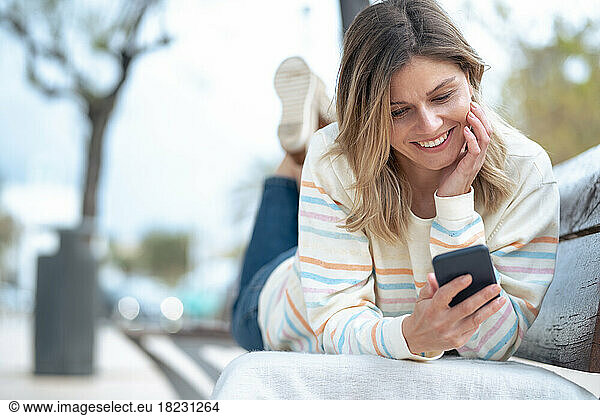 Smiling young woman lying on bench using mobile phone