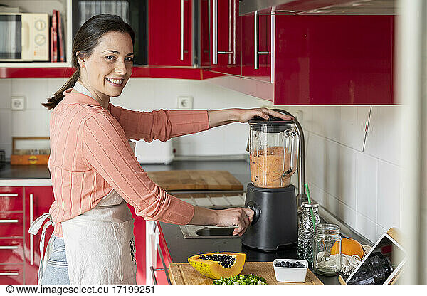 Smiling young woman preparing healthy fruit smoothie in kitchen at home