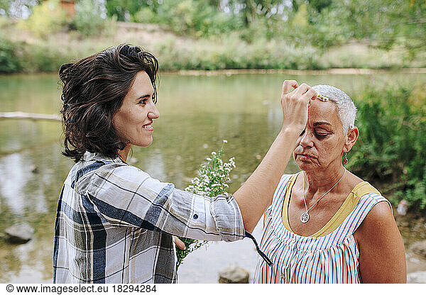 Smiling young woman putting flower on friend's head by lake in park