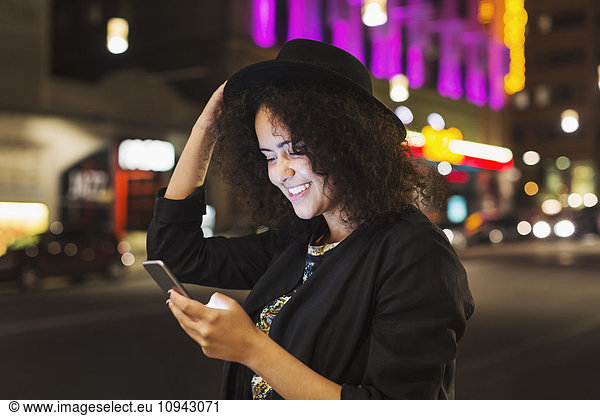 Smiling young woman reading text messaging on smart phone in city at night