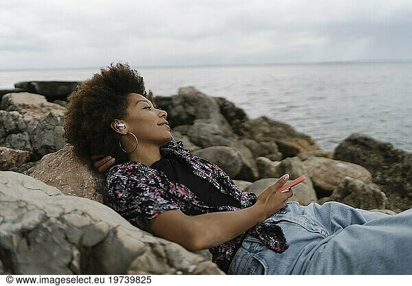 Smiling young woman relaxing on rocks and listening to music through wireless In-ear headphones