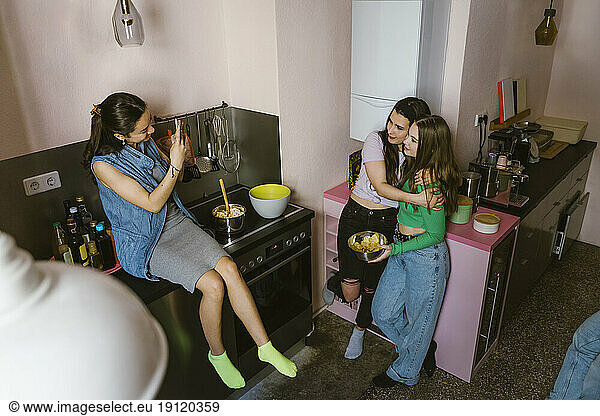 Smiling young woman sitting on counter photographing female friends in kitchen at home