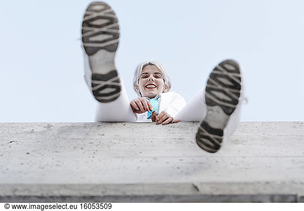 Smiling young woman sitting on retaining wall against clear sky in city