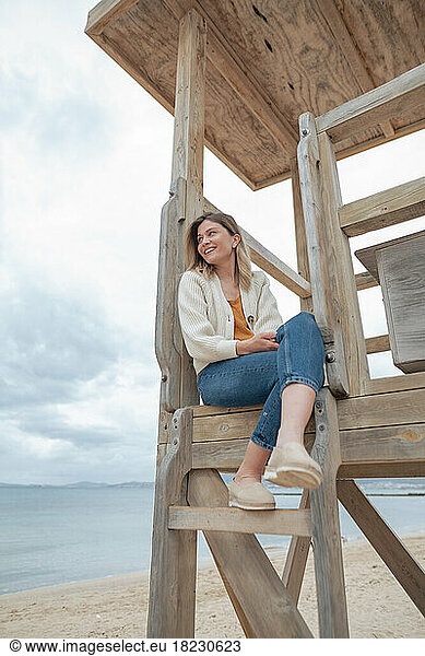 Smiling young woman sitting with legs crossed on wooden lifeguard hut at beach