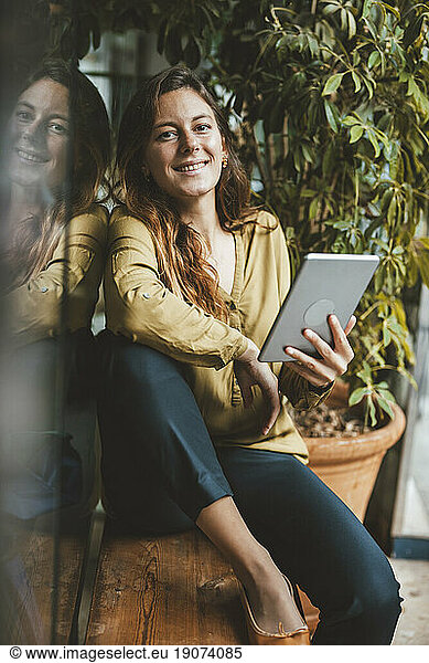 Smiling young woman sitting with tablet PC on seat in cafe