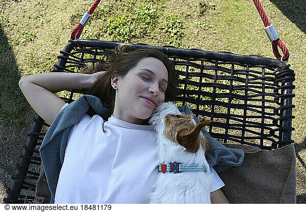 Smiling young woman sleeping with dog on hammock at park