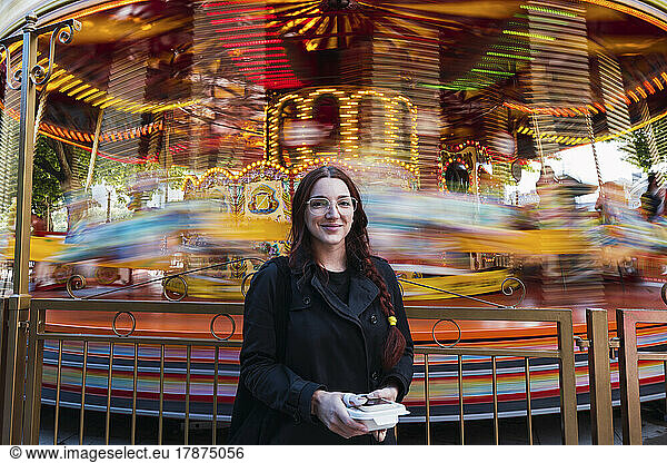 Smiling young woman standing in front of carousel at amusement park