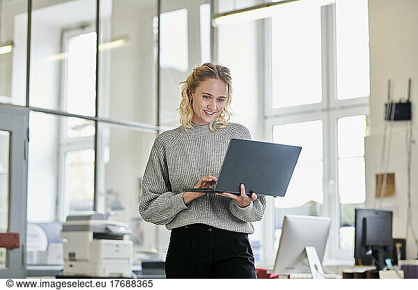 Smiling young woman standing in office using laptop