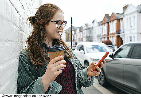 Smiling young woman standing with disposable coffee cup and using smart phone