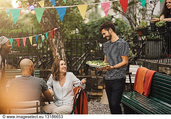 Smiling young woman talking with male friend carrying food during garden party
