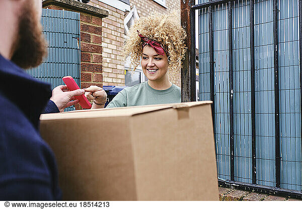 Smiling young woman touching smart phone held by delivery person