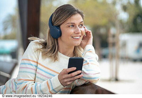 Smiling young woman using smart phone listening to music sitting on bench